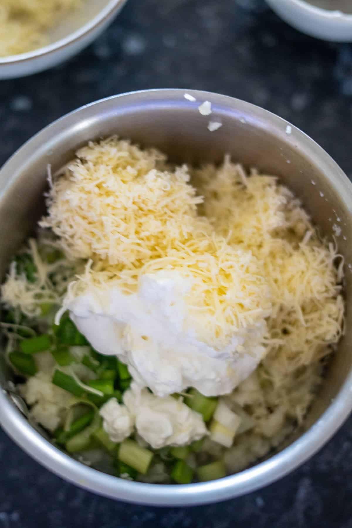 A bowl featuring toppings for a savory low carb cauliflower bake: shredded cheese, chopped green onions, and a dollop of cream rest elegantly on a dark countertop.