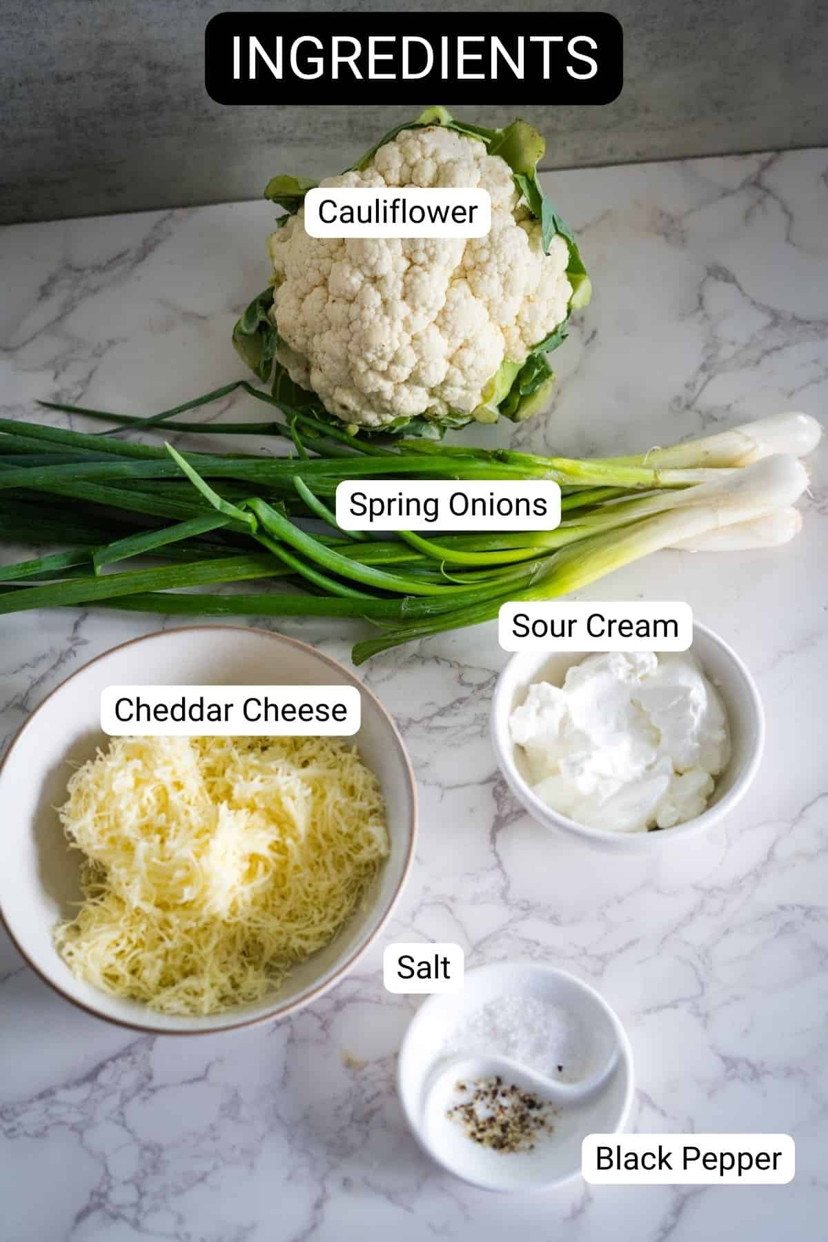 Ingredients for a low carb cauliflower bake on a marble surface: cauliflower, spring onions, cheddar cheese, sour cream, salt, and black pepper.