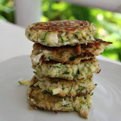 A stack of five keto zucchini fritters on a white plate, featuring visible chunks of zucchini and a crispy, golden-brown exterior.