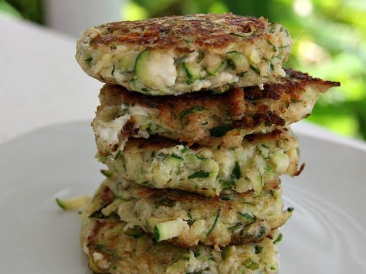 A stack of five keto zucchini fritters on a white plate, featuring visible chunks of zucchini and a crispy, golden-brown exterior.