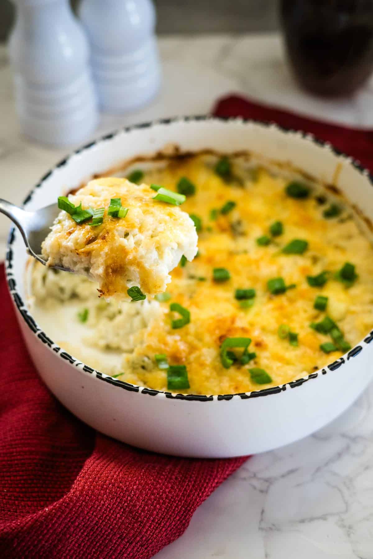 A delicious dish of low carb cauliflower bake, topped with melted cheese and chopped green onions. A spoon lifts out a portion, revealing the creamy layers beneath.