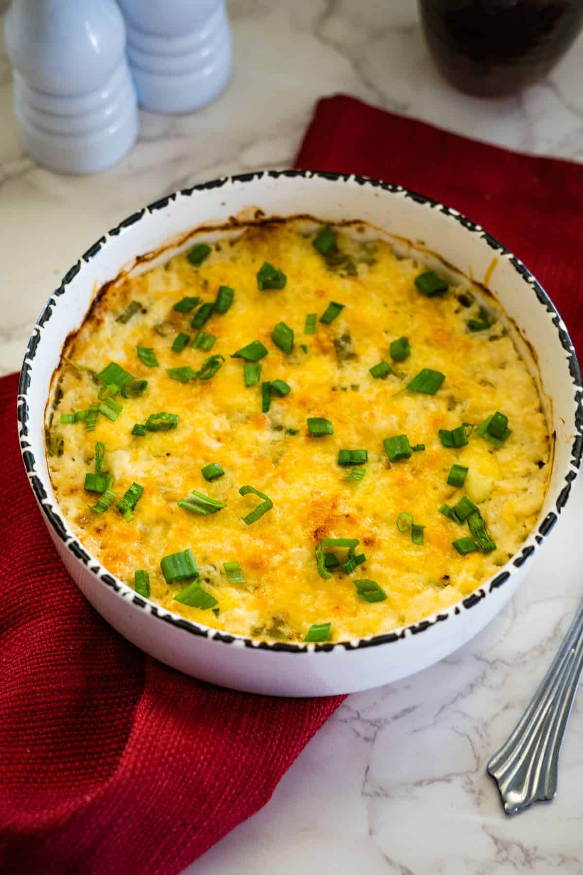 A delicious low carb cauliflower bake featuring a cheesy casserole topped with chopped green onions, all nestled in a white circular dish on a red cloth.