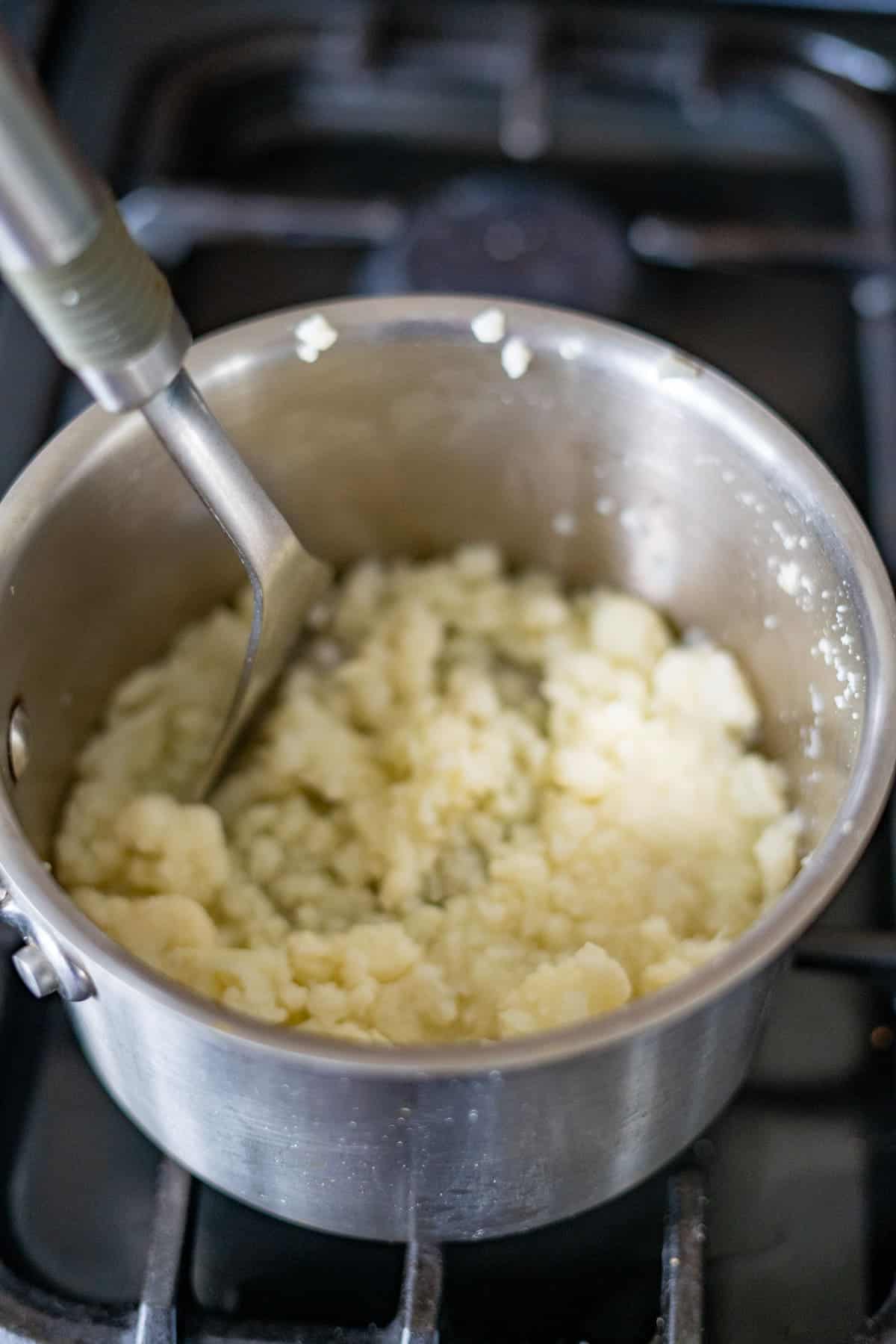 A low carb twist on a classic, creamy mashed potatoes are being stirred in a silver pot on the stovetop, reminiscent of making a delicious cauliflower bake.