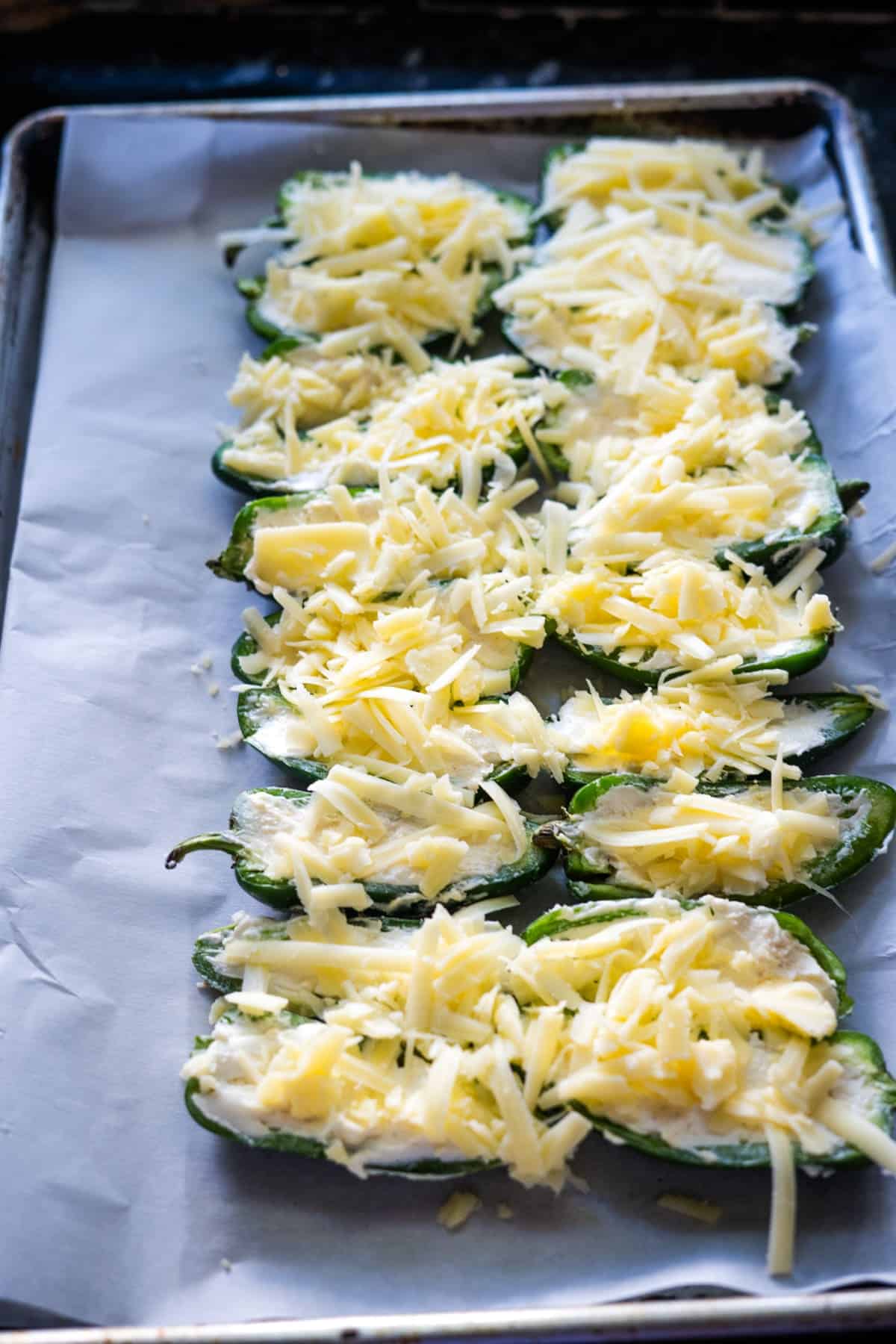 Stuffed jalapeño halves on a baking sheet are generously topped with shredded cheese, ready for baking.