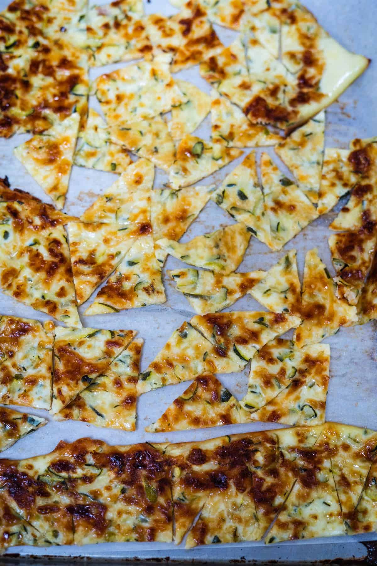 A baking sheet with a batch of keto flatbread that has been sliced into various shapes and sizes after baking, featuring a golden-brown, cheesy top reminiscent of zucchini nachos.