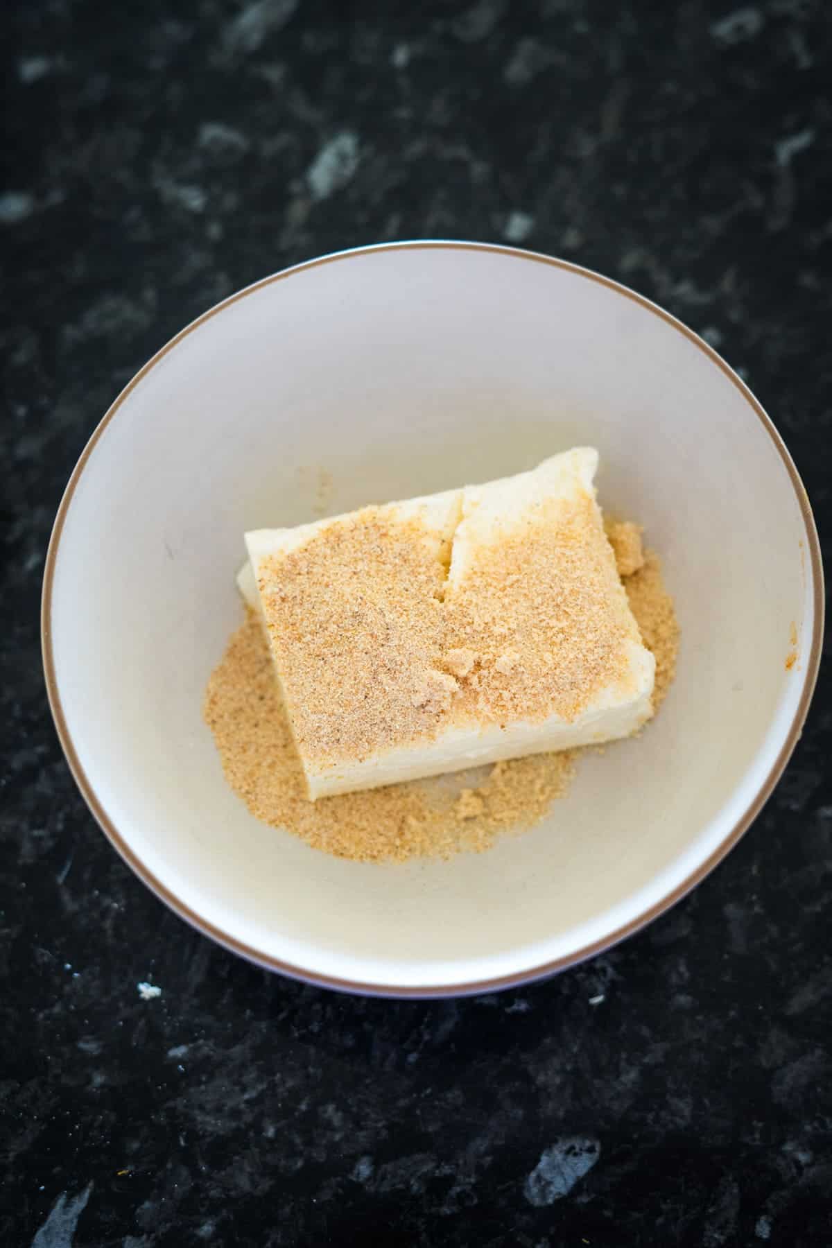 A bowl contains a block of butter partially covered in brown sugar, nestled on a dark marble countertop next to a plate of stuffed jalapenos.