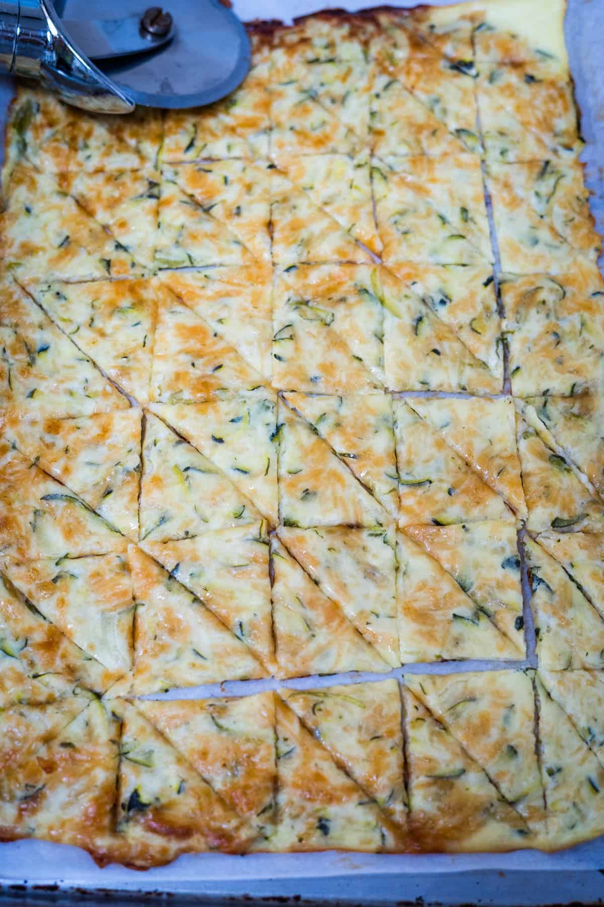 A rectangular cheese and vegetable dish, resembling keto zucchini nachos, cut into a grid pattern with some triangles, partially sliced with a pizza cutter on the side.