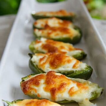 A row of stuffed jalapeño poppers filled with melted cheese lines a white rectangular plate.