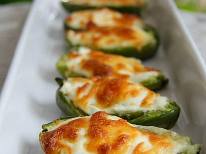 A row of stuffed jalapeño poppers filled with melted cheese lines a white rectangular plate.