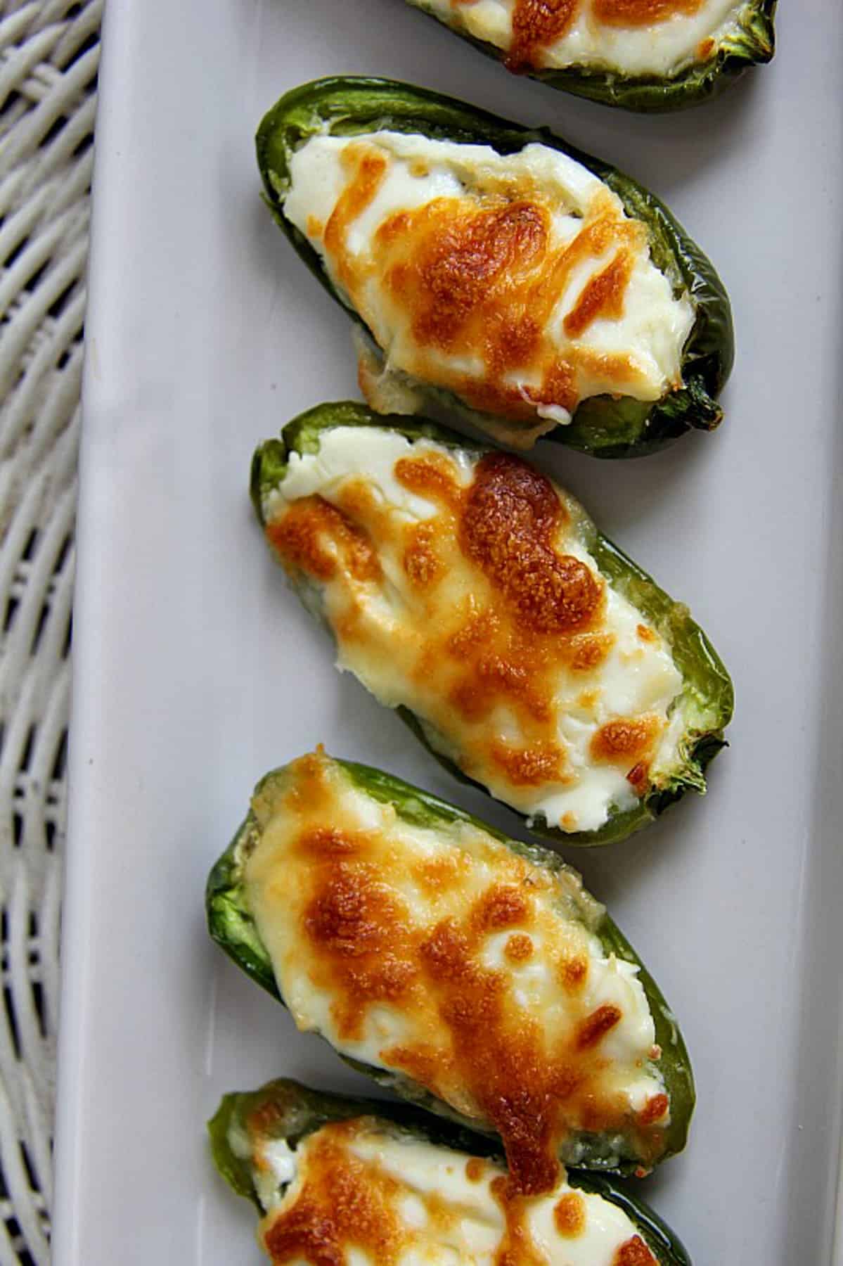 Five stuffed jalapeño peppers brimming with melted cheese and perfectly browned on top, arranged in a row on a white plate.