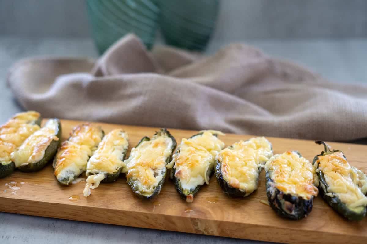 Eight stuffed jalapeño peppers bursting with cheese rest on a wooden board, their tops gloriously melted. A textured cloth provides the perfect backdrop.