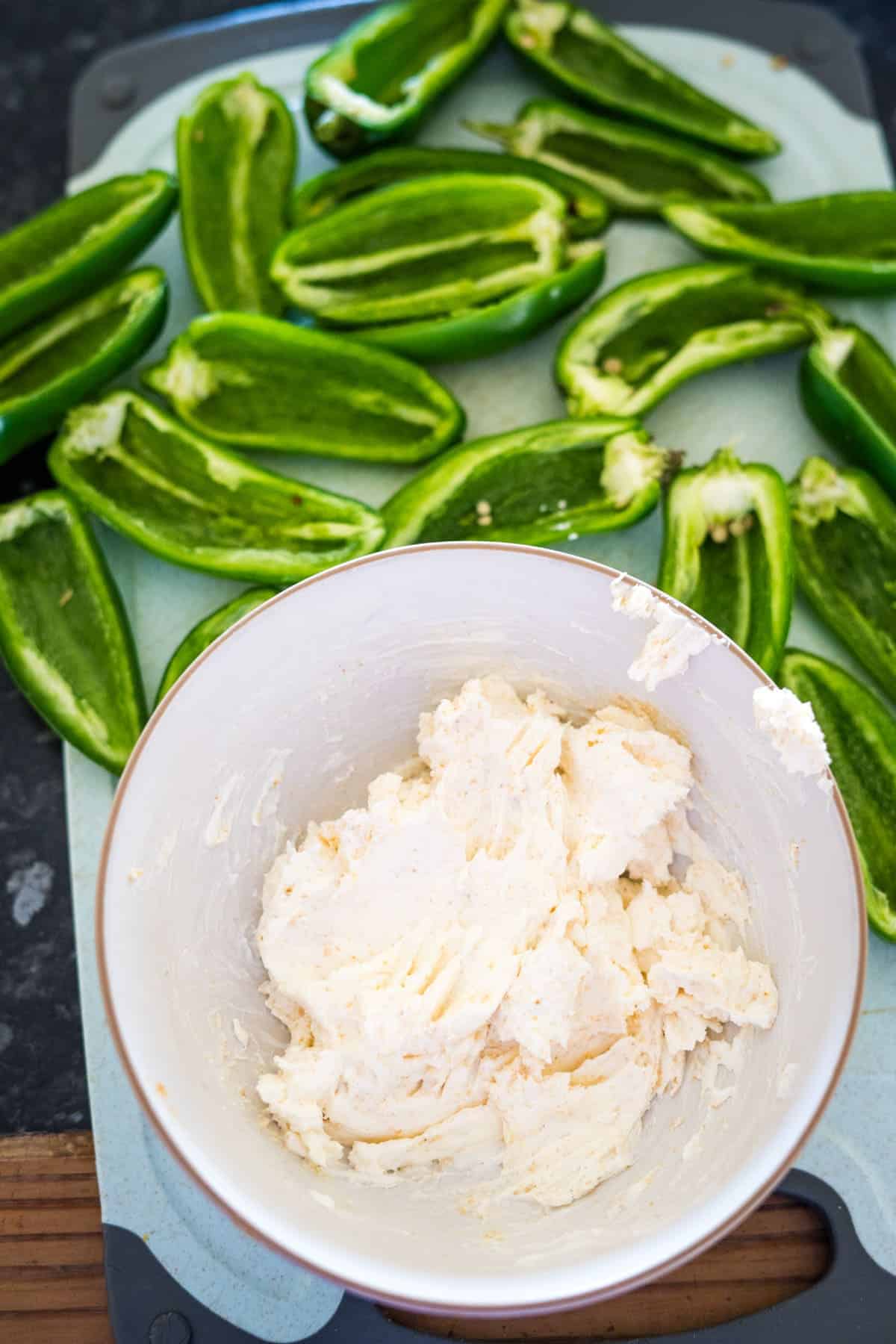 Halved green jalapeños on a cutting board await their transformation into delicious stuffed jalapeños, next to a bowl of creamy cheese filling.