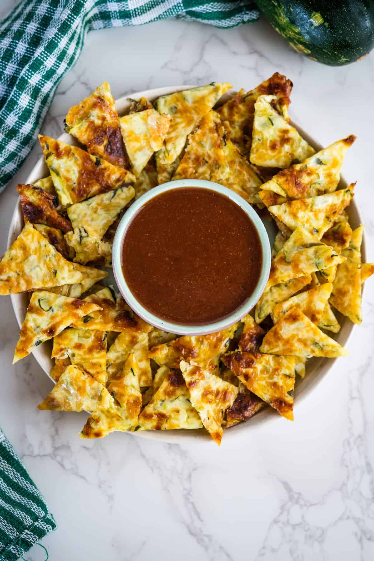 A plate of triangular cheese crisps arranged around a bowl of brown dipping sauce, placed on a marble surface with a green checkered cloth partially visible, offers the perfect setup for keto zucchini nachos.