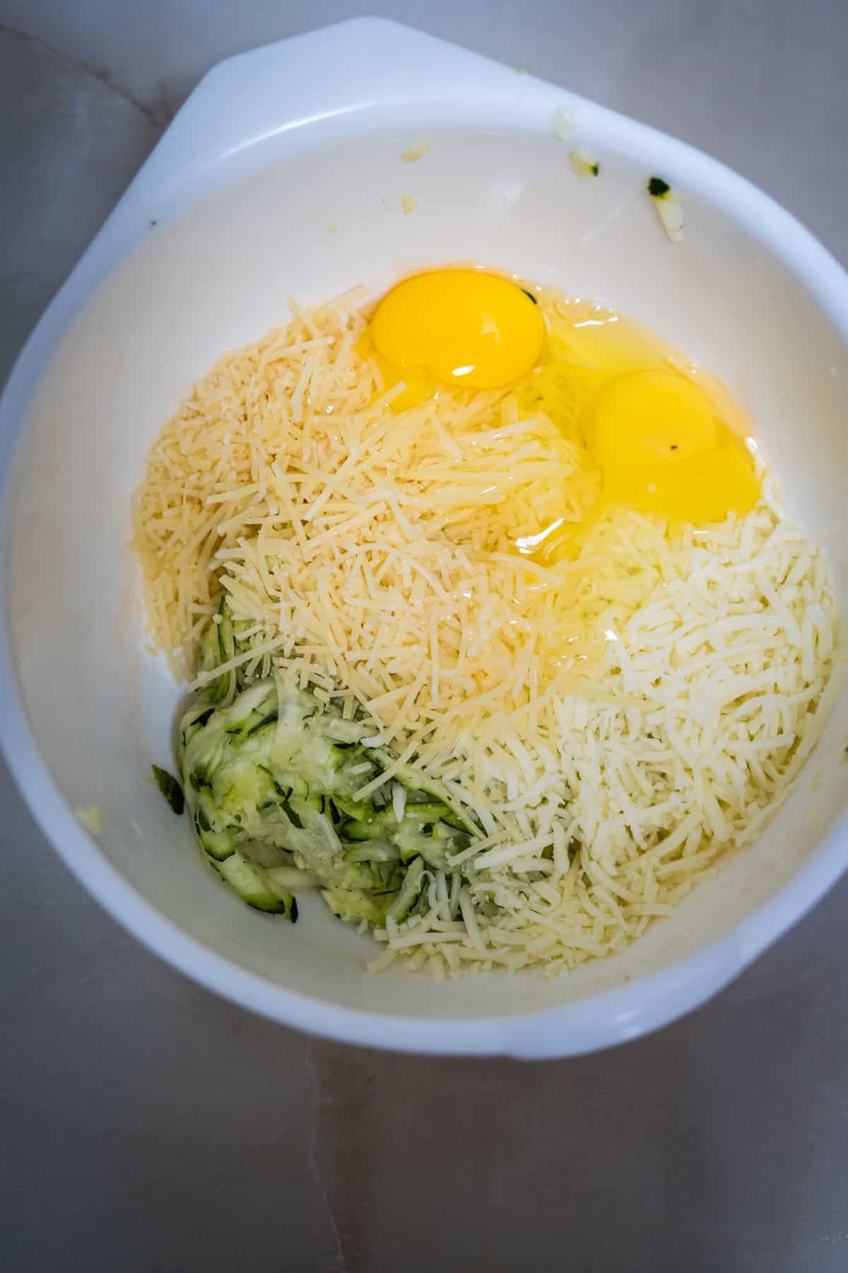 A white mixing bowl containing two cracked eggs, shredded cheese, and finely chopped green vegetables for keto zucchini nachos placed on a smooth surface.