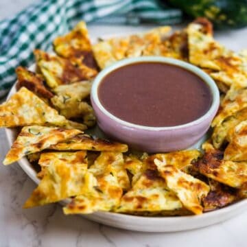 A platter of sliced cheesy garlic zucchini bread arranged in a circle around a bowl filled with marinara sauce, reminiscent of keto zucchini nachos.