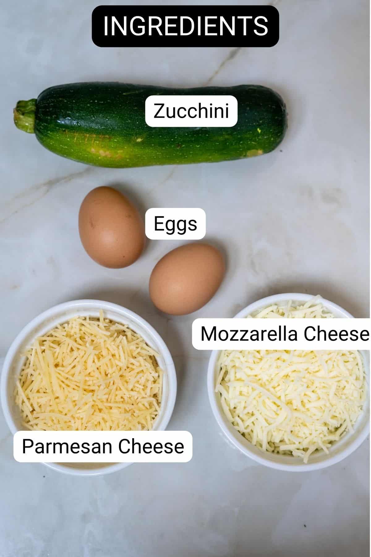 Ingredients displayed on a table for keto zucchini nachos: a zucchini, two eggs, a bowl of shredded parmesan cheese, and a bowl of shredded mozzarella cheese. Each item is labeled.