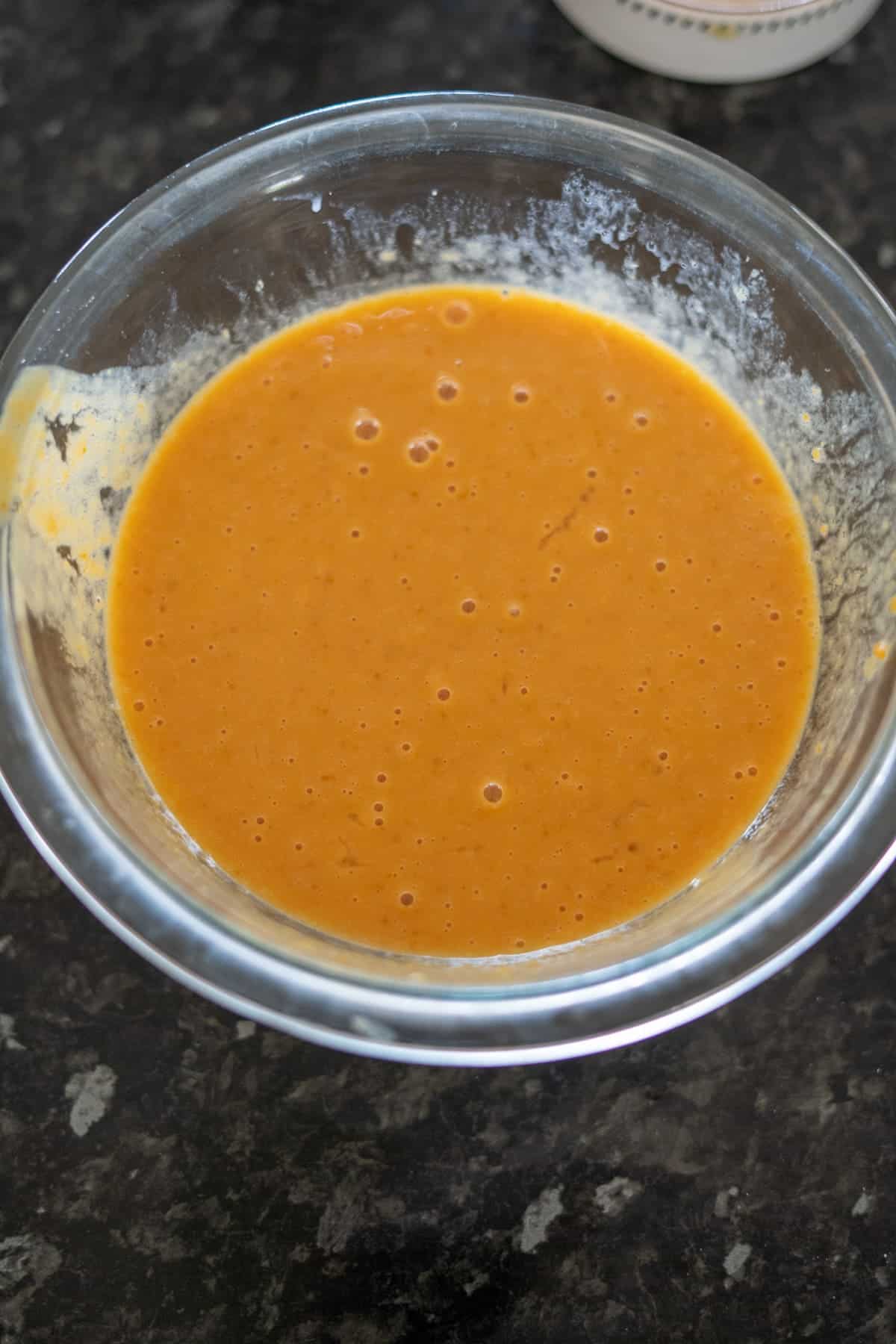 A glass bowl containing orange-colored keto pumpkin bread batter rests on a dark countertop.