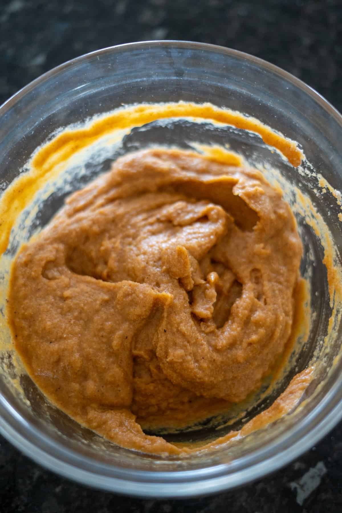 A glass bowl sits on the dark countertop, filled with smooth, orange-brown keto pumpkin bread batter.