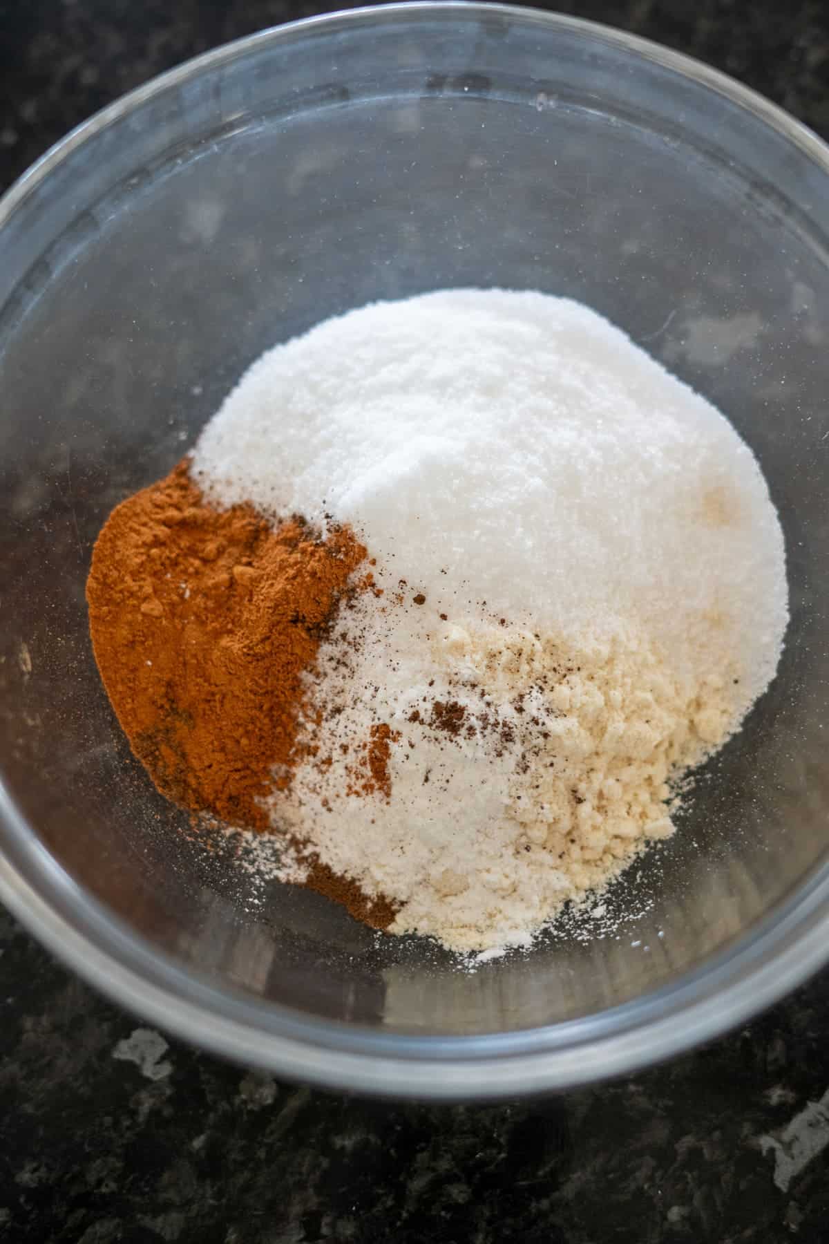 A glass bowl containing sugar, cinnamon, and flour sits on a dark countertop, ready to transform into delicious keto pumpkin bread.