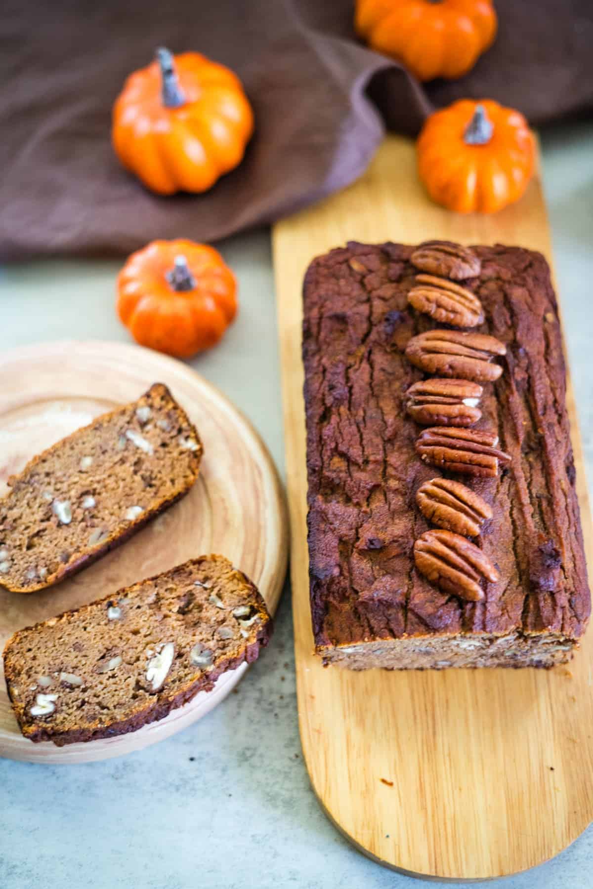 A sliced loaf of keto pumpkin bread topped with pecans rests on a wooden board, surrounded by small decorative pumpkins.