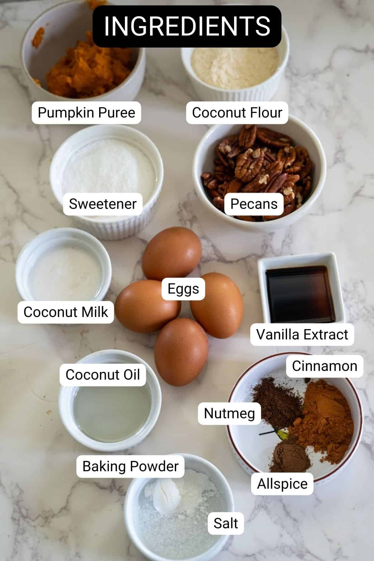 Ingredients for keto pumpkin bread on a marble counter include pumpkin puree, coconut flour, sweetener, pecans, coconut milk, eggs, vanilla extract, coconut oil, nutmeg, cinnamon, allspice, baking powder, and salt.