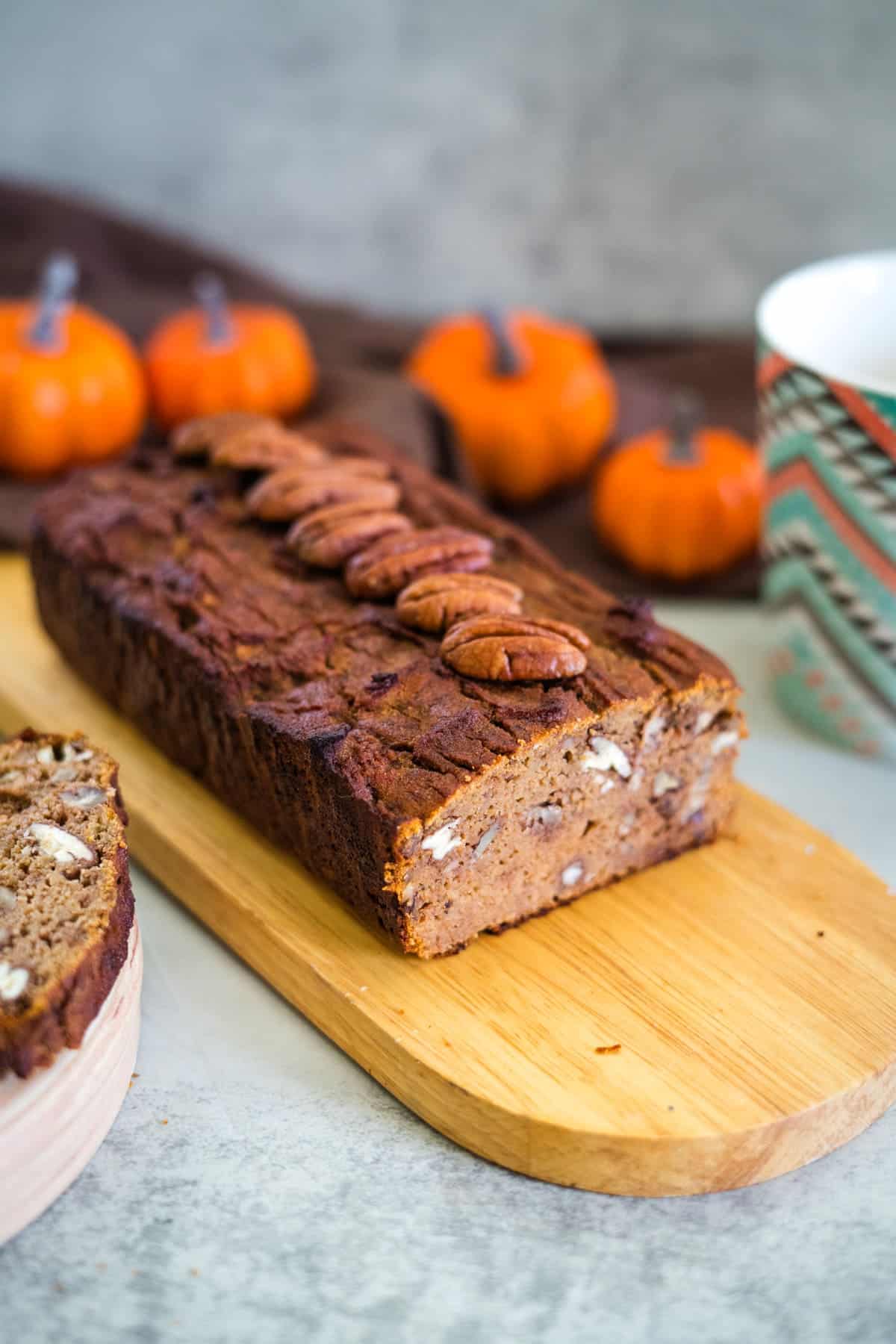 A loaf of keto pumpkin bread with pecans on top sits on a wooden board. Several small pumpkins, a ceramic mug, and a brown cloth create a cozy autumn backdrop.