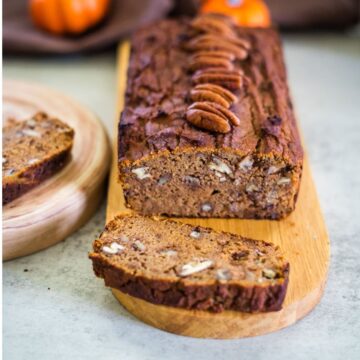 A loaf of keto pumpkin bread sits on a wooden board, topped with whole pecans. Two slices are cut, with one resting invitingly on a nearby plate.