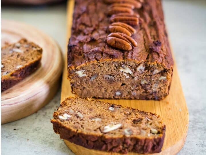 A loaf of keto pumpkin bread sits on a wooden board, topped with whole pecans. Two slices are cut, with one resting invitingly on a nearby plate.