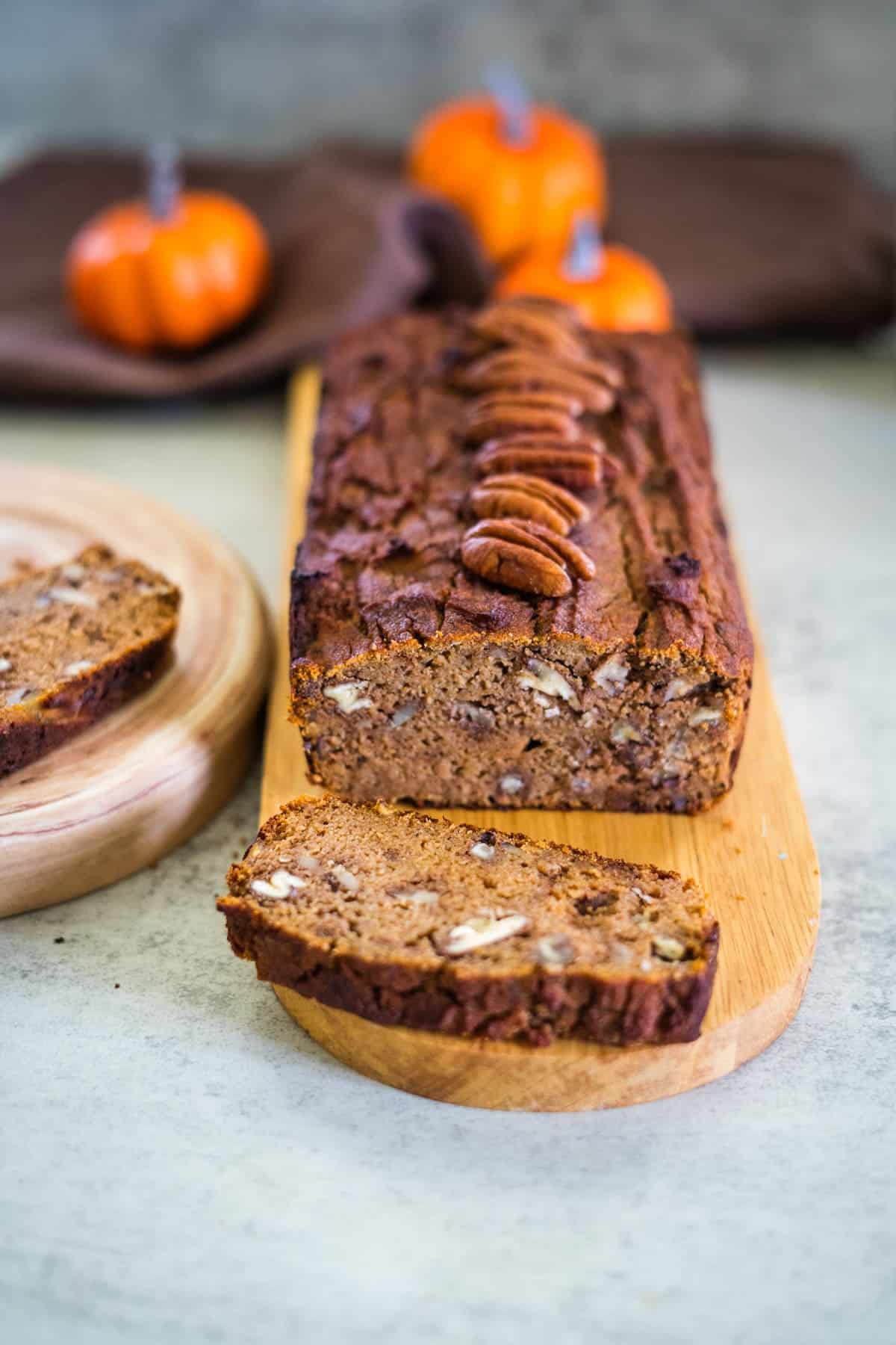 A loaf of keto pumpkin bread topped with pecans rests on a wooden board, surrounded by whole pumpkins in the background.