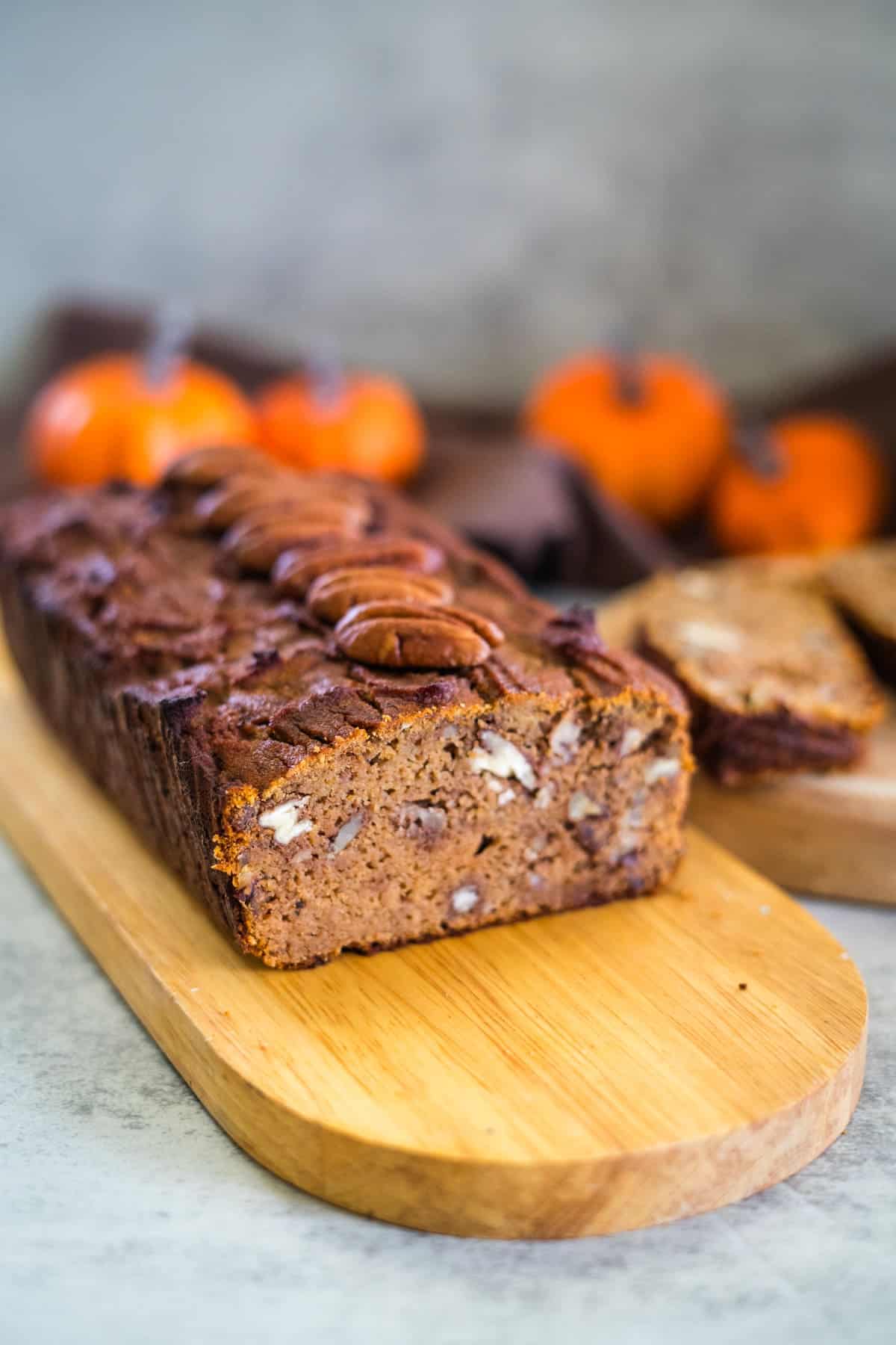 A loaf of keto pumpkin bread with pecans rests on a wooden board, surrounded by slices of the treat and small pumpkins in the background.