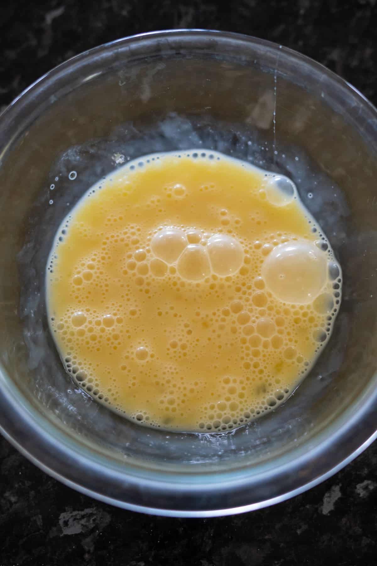 A clear glass bowl containing beaten eggs with bubbles on the surface sits on a dark countertop, ready to be transformed into delicious keto pumpkin bread.