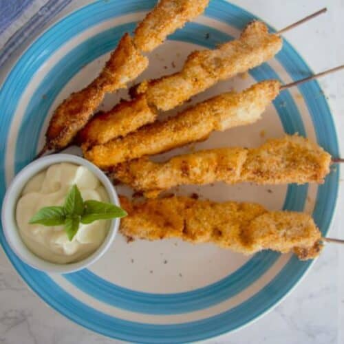 A blue-rimmed plate holds five skewered coconut chicken pieces next to a small white bowl of creamy dipping sauce garnished with basil leaves.