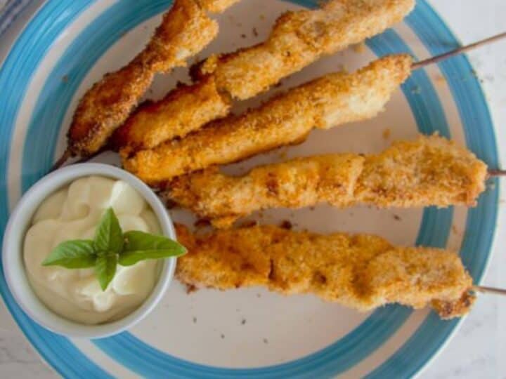 A blue-rimmed plate holds five skewered coconut chicken pieces next to a small white bowl of creamy dipping sauce garnished with basil leaves.