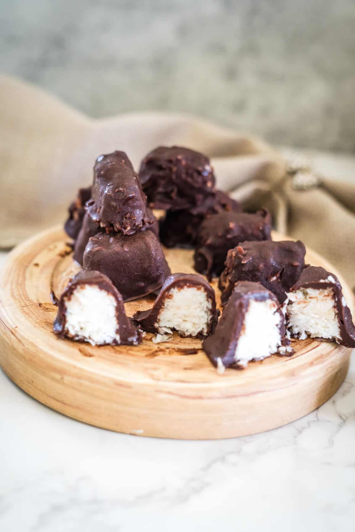 Wooden plate with chocolate-covered bounty bars, some sliced to reveal the white coconut filling, on a light marble surface with a tan cloth in the background.