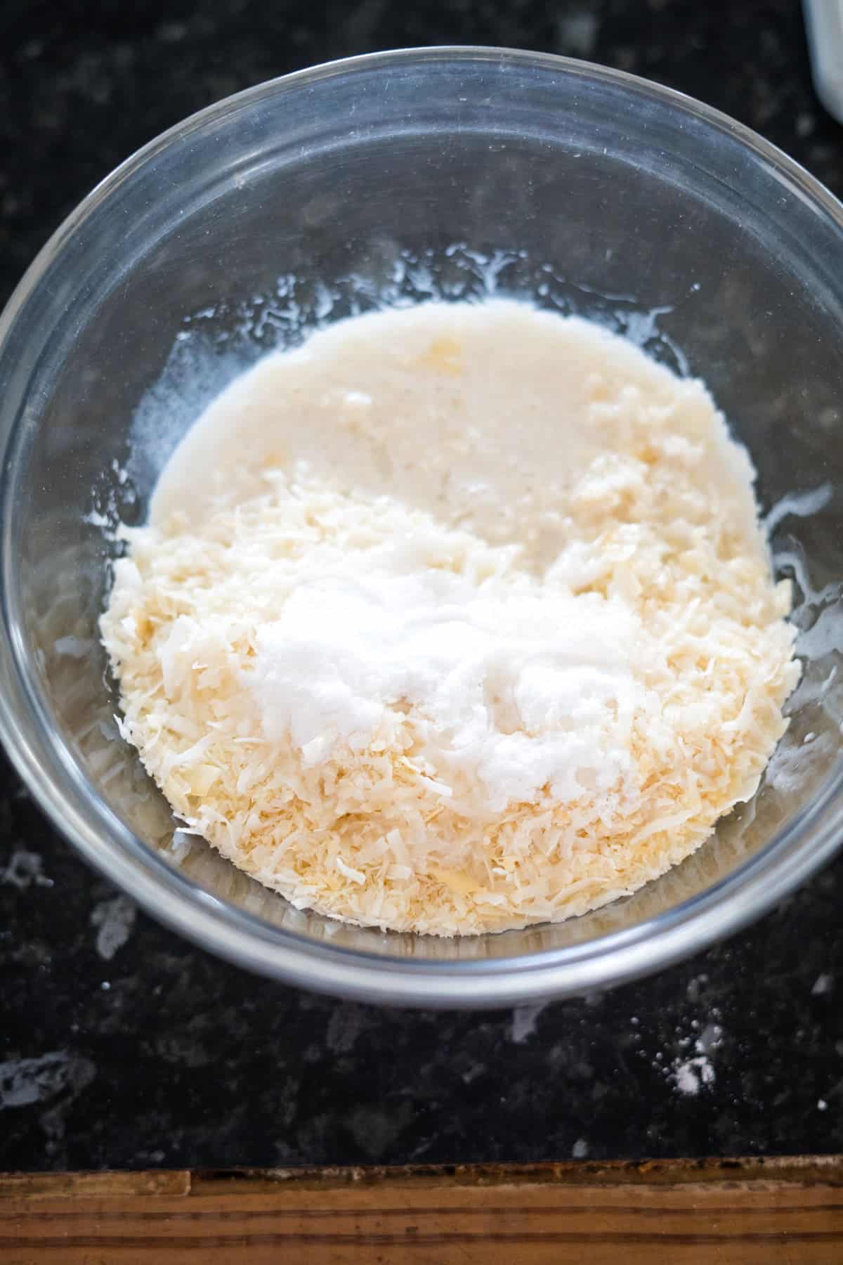 A glass bowl on a dark countertop holds a delightful mixture reminiscent of bounty bars, with grated coconut, sugar, and a liquid ingredient melding together.