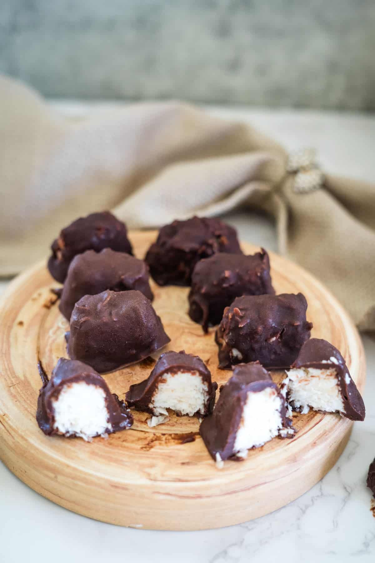 A wooden platter showcases bounty bars, with chocolate-covered coconut treats. One is cut in half, revealing the luscious filling. A beige cloth elegantly rests in the background.