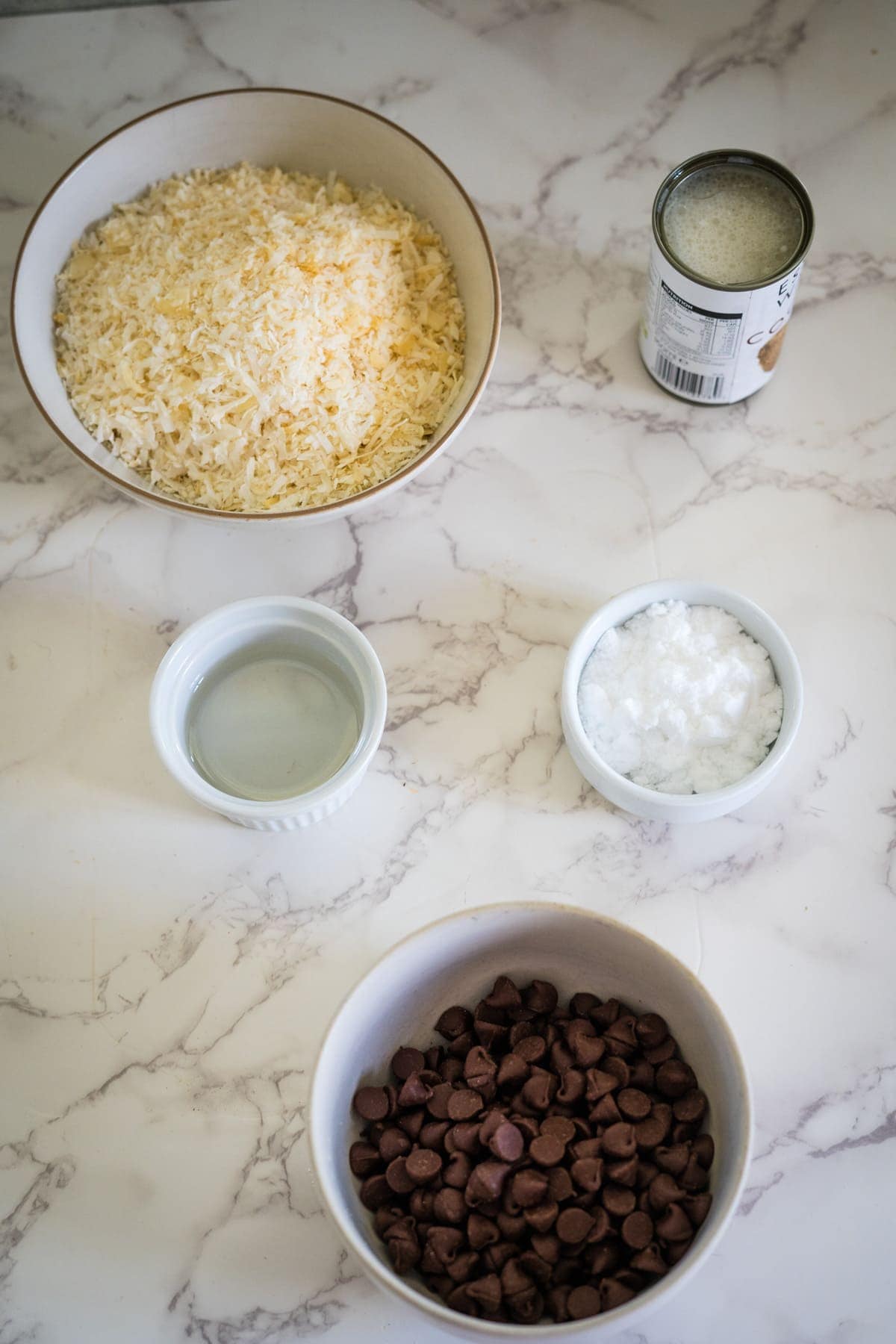 Ingredients for homemade bounty bars on a marble surface: shredded coconut in a bowl, a can of condensed milk, a small bowl of oil, a bowl of coconut flakes, and a bowl of chocolate chips.