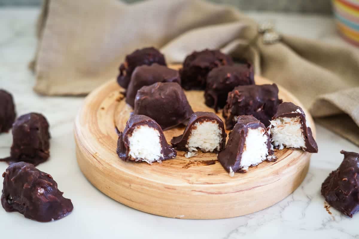 A wooden platter with chocolate-covered coconut candies, reminiscent of homemade bounty bars, some whole and some cut in half, rests elegantly on a marble surface with a beige cloth in the background.