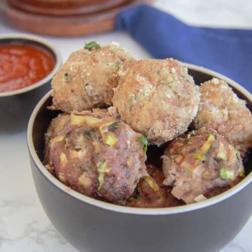 A bowl of homemade chicken meatballs with visible herbs, accompanied by a side of tomato sauce on a marble countertop.