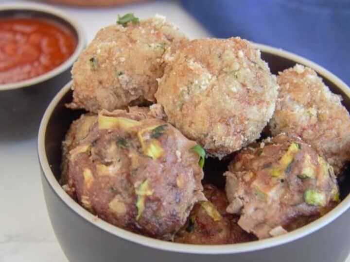 A bowl of homemade chicken meatballs with visible herbs, accompanied by a side of tomato sauce on a marble countertop.