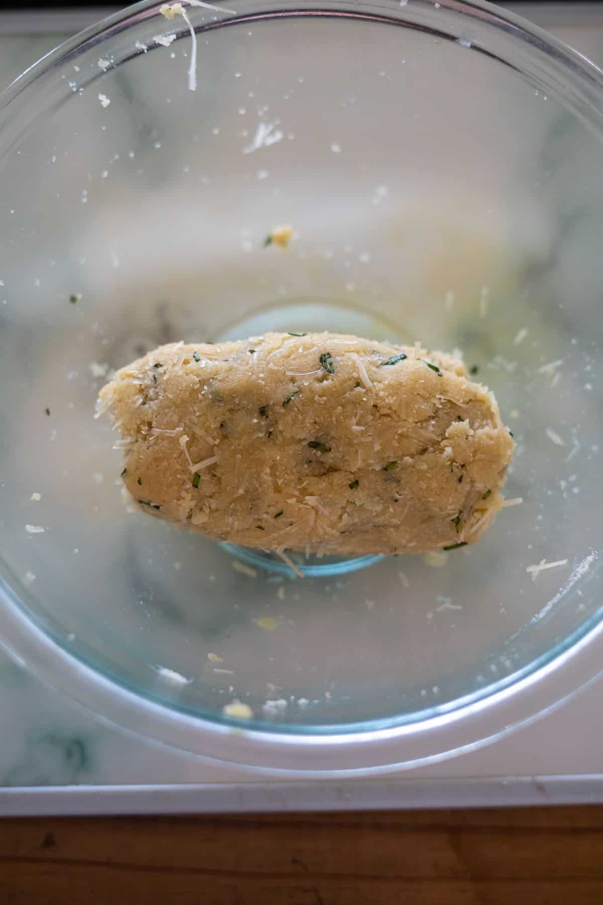 A glass bowl contains a mixture of dough infused with scattered rosemary and Parmesan cheese.