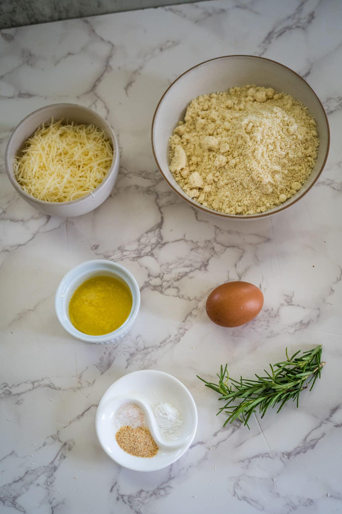 Ingredients on a marble surface: grated rosemary parmesan cheese, almond flour, melted butter, an egg, fresh rosemary, garlic powder, and baking powder in separate bowls.