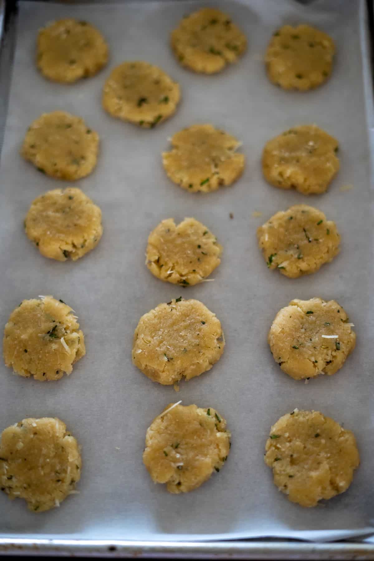 Rosemary-infused cookie dough rounds, enriched with a hint of parmesan, are artfully arranged on a baking sheet lined with parchment paper.