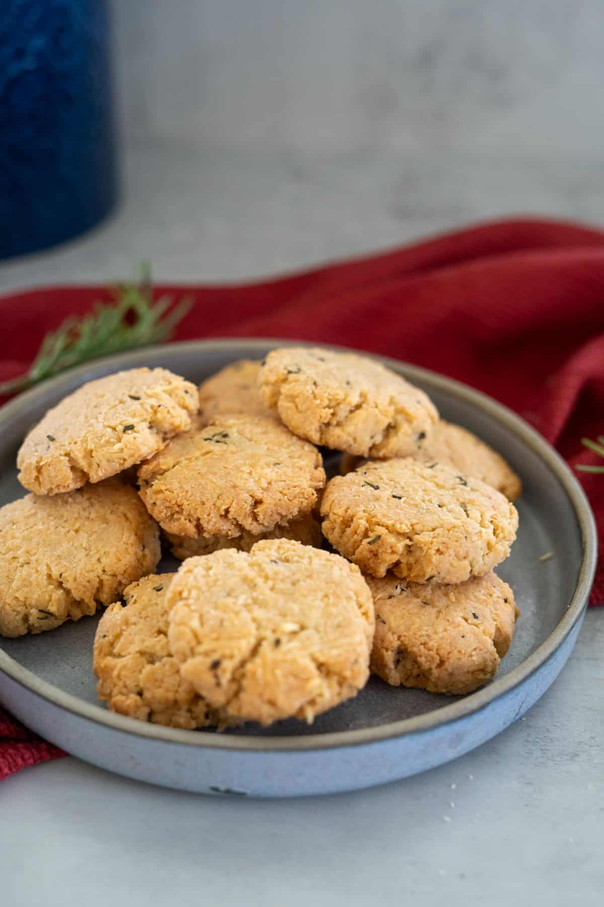 A plate of round, golden-brown rosemary parmesan cookies rests on a light gray surface, accented by a vibrant red cloth in the background.