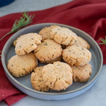 A gray plate holds a pile of round, homemade rosemary-parmesan cookies on a red cloth. Sprigs of rosemary are artfully placed nearby.