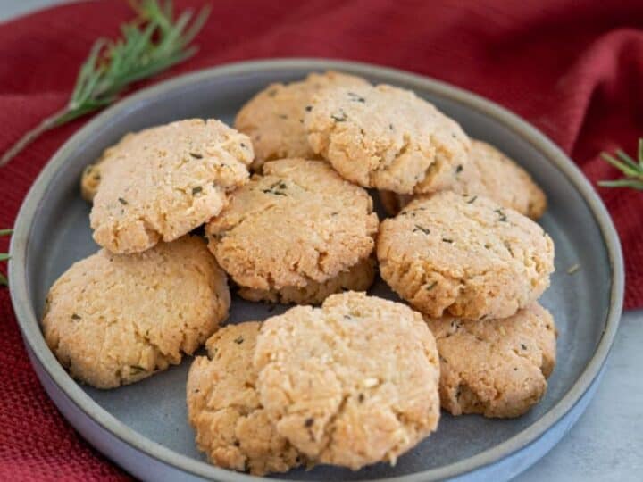 A gray plate holds a pile of round, homemade rosemary-parmesan cookies on a red cloth. Sprigs of rosemary are artfully placed nearby.