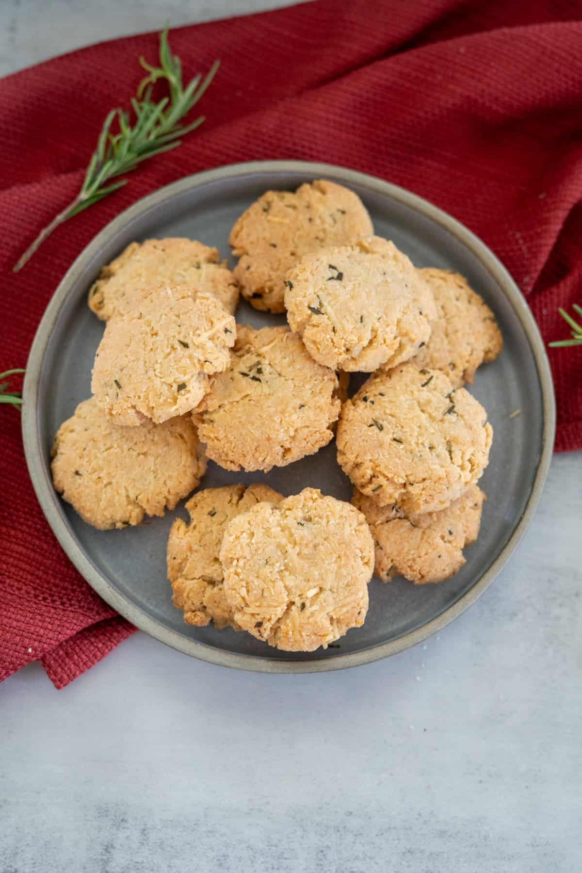 A plate of homemade rosemary parmesan cookies rests on a gray surface, accented by a red cloth and sprigs of fresh rosemary nearby.