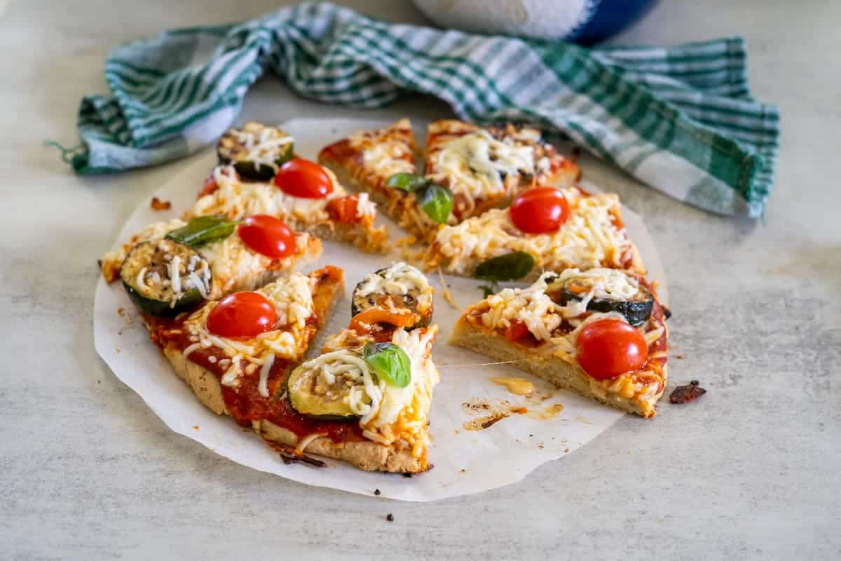 A sliced vegetable pizza made with almond flour, topped with tomatoes and basil on parchment paper, alongside a green checkered cloth.