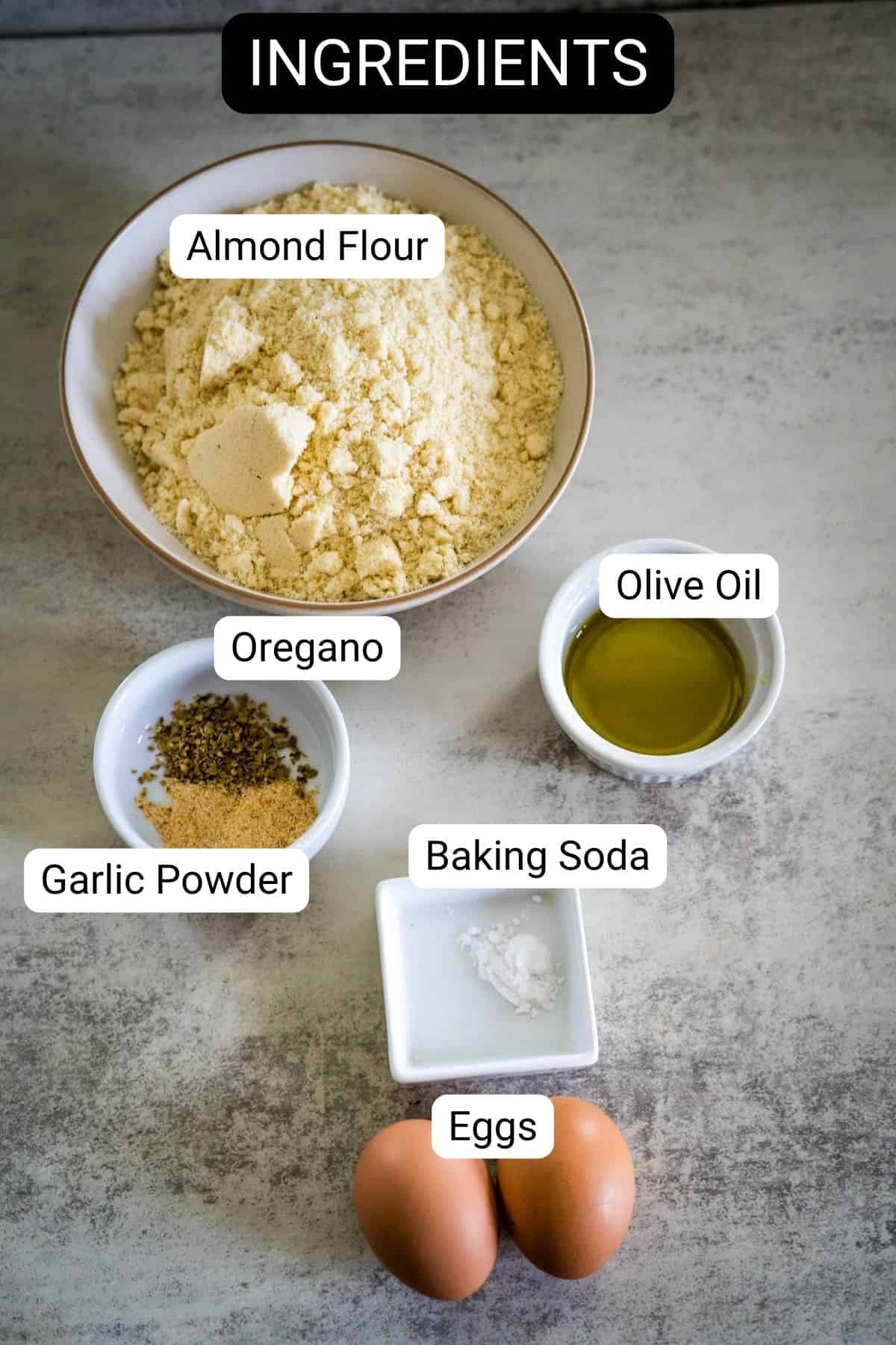 A bowl of almond flour sits alongside a cup of olive oil, while oregano, garlic powder, and baking soda add a dash of flavor. Two eggs complete the inviting array on the table.