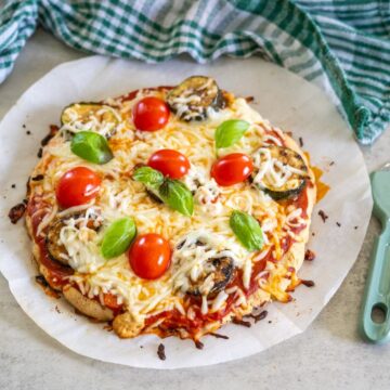 Homemade pizza with melted cheese, cherry tomatoes, basil leaves, and grilled zucchini slices sits on parchment paper made with almond flour crust, nestled beside a green plaid cloth.